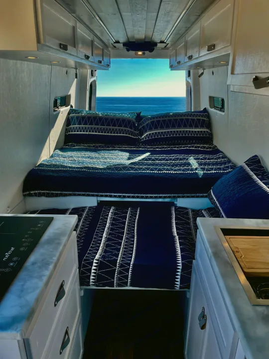 Step inside this RV rental! A comfy bed with blue bedding faces stunning ocean views, with a compact kitchen in the foreground.. Mercedes-Benz Sprinter 2022