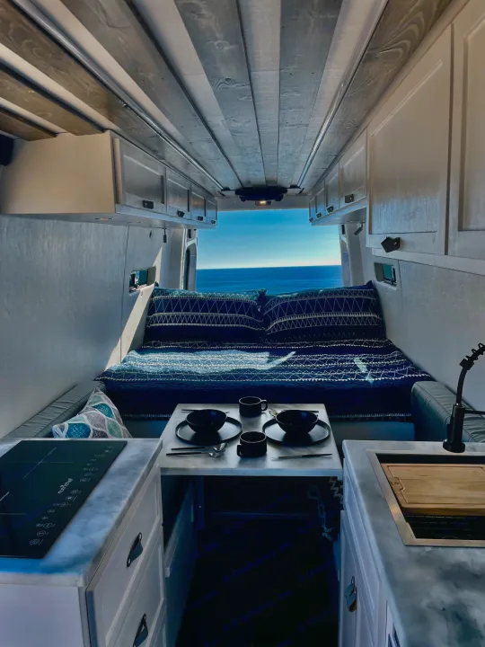Cozy RV rental interior with ocean view bed, kitchen, and dining area. Perfect for van life adventures.. Mercedes-Benz Sprinter 2022