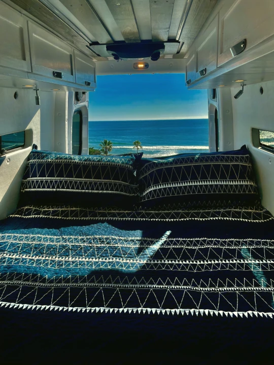 Inside an **rv rental**, a comfy bed offers stunning ocean views and distant palm trees. Ideal for a serene camper van getaway.. Mercedes-Benz Sprinter 2022
