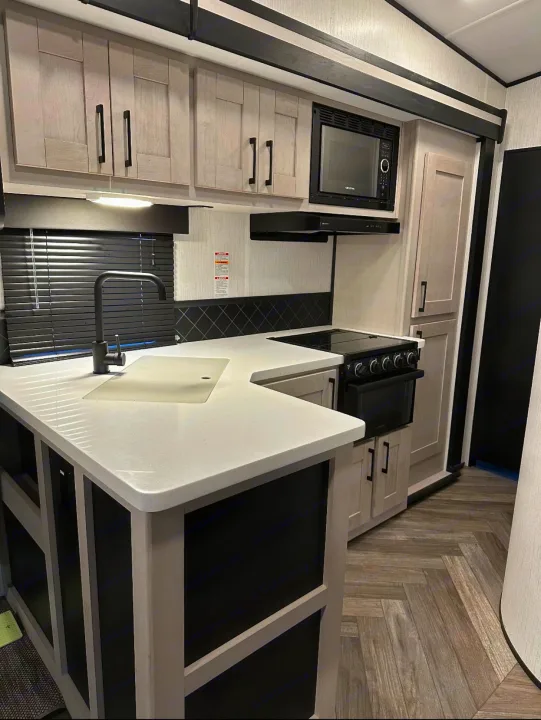 Inside a modern RV rental, this kitchen features light gray cabinets, white counters, black appliances, and herringbone flooring.. Heartland Torque 2022