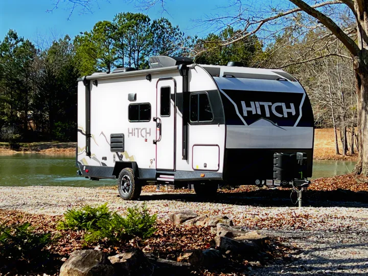 A sleek 'HITCH' travel trailer, perfect for an RV rental adventure, parked by a scenic lake amidst trees.. Cruiser Rv Corp Hitch 2022