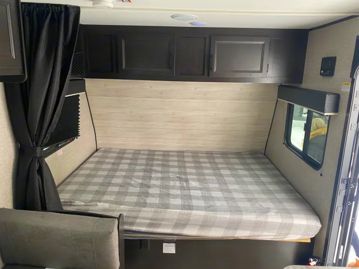 Bright RV rental interior featuring a large plaid bed, dark overhead cabinets, and a side window, offering a comfy sleeping space.. Jayco Jay Flight SLX STX Edition 2022