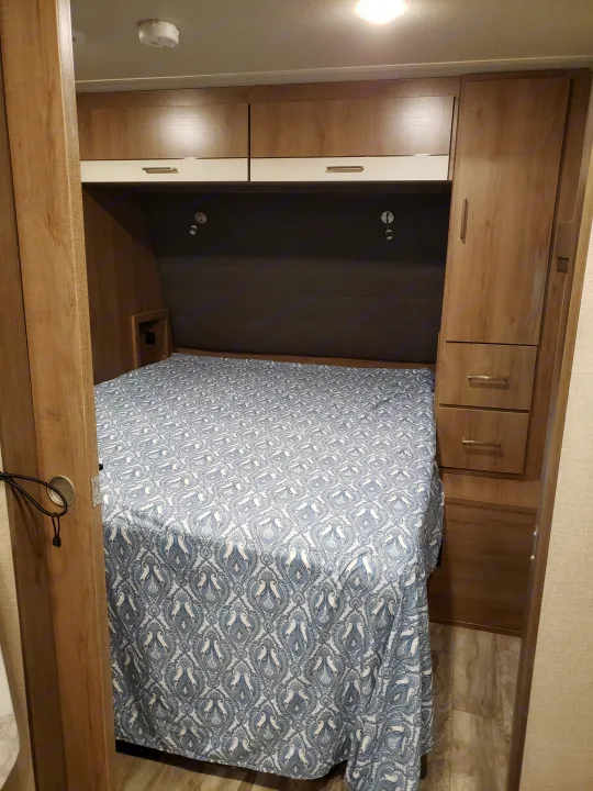 Inside an RV rental: a compact bedroom with a patterned bedspread, overhead cabinets, and reading lights.. Grand Design Imagine XLS 2022