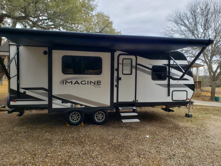 This sleek white and black RV rental, with its awning extended, awaits your next outdoor adventure on a gravel lot.. Grand Design Imagine XLS 2022