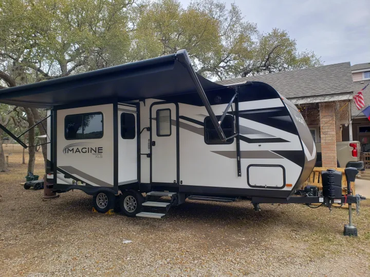 This white & black Imagine XLS travel trailer with an extended awning is parked on gravel, an excellent RV rental for your next trip.. Grand Design Imagine XLS 2022