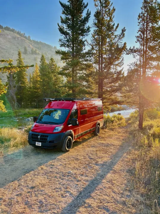Red RV rental parked by a river, nestled among sunlit trees & mountains on a dirt path. Your perfect outdoor escape awaits.. Dodge promaster 3500 2018