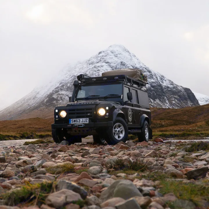 Rugged Land Rover Defender with roof tent on rocky terrain, snowy mountain backdrop. Your next adventure rv rental awaits!. Land Rover Defender 110 XS Utility + 2 Roof Tents 2013