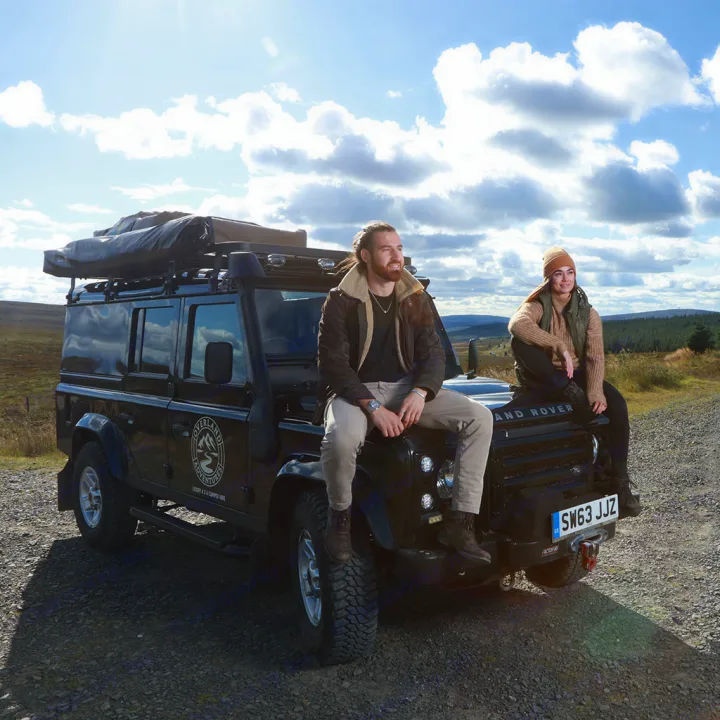 Explore new horizons! Two people on a Land Rover Defender overland RV rental, ready for adventure & camping in a vast landscape.. Land Rover Defender 110 XS Utility + 2 Roof Tents 2013