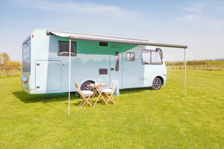 Light blue RV rental with an extended awning, outdoor dining table, and chairs set up on a sunny, grassy field.. GlamperRV DBL 2026