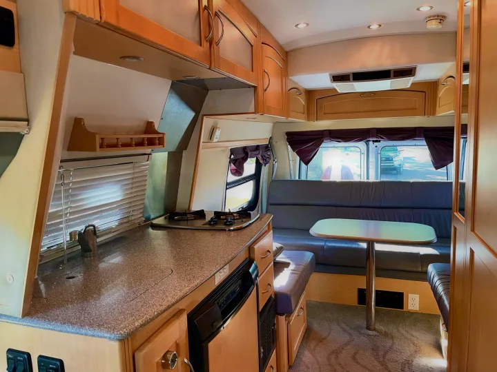 Interior of an RV rental showing a kitchen counter with stove/sink, wood cabinets, and a dining area with bench seating.. Pleasure Way Excel 2007