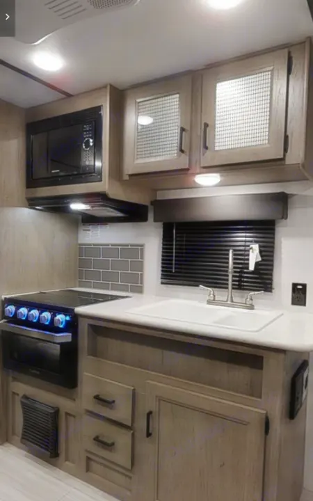 An RV rental kitchen, featuring a built-in microwave, stove, sink, and light wood cabinetry.. Keystone Hideout 21BHWE 2021