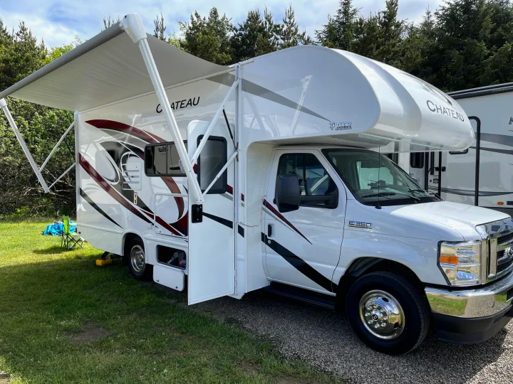 White motorhome RV rental, awning out, parked on a grass and gravel campsite.. Thor Chateau 2022