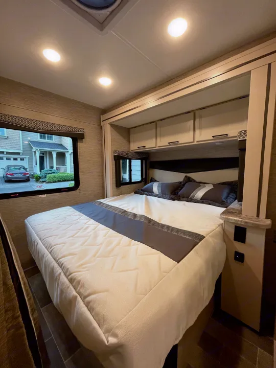Cozy RV rental bedroom showcasing a bed with gray and white bedding, built-in cabinets, and a window view for comfortable travel.. Thor Chateau 2022