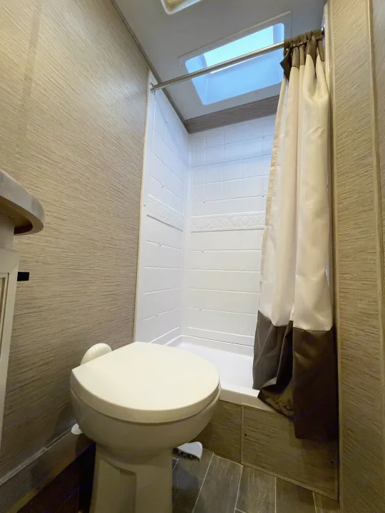RV rental bathroom features a compact shower with white base, patterned walls, and curtain, alongside a toilet. Light wood-look floor.. Thor Chateau 2022