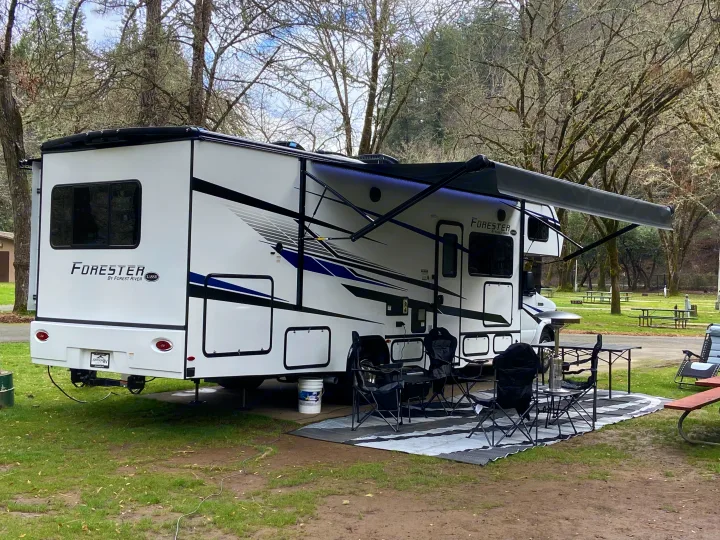 This white RV rental is perfectly set up at a treed campsite with an extended awning and comfy outdoor living space.. Forest River Forester 2023
