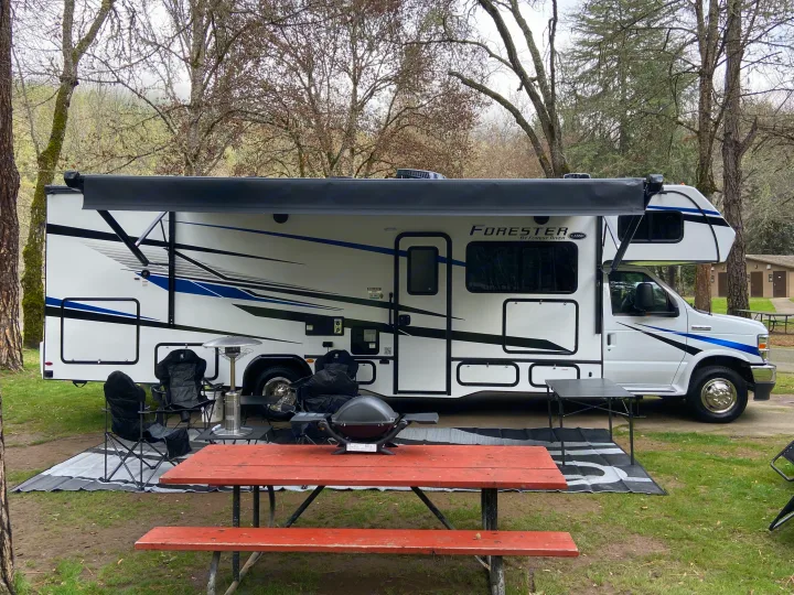 This RV rental is parked at a serene campsite, complete with a picnic table, grill, chairs, and awning for outdoor leisure.. Forest River Forester 2023