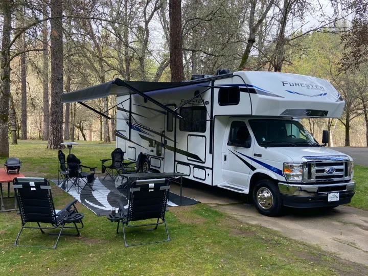 This rv rental is set up for comfortable camping, featuring an extended awning, outdoor chairs, and a grill at a treed campsite.. Forest River Forester 2023