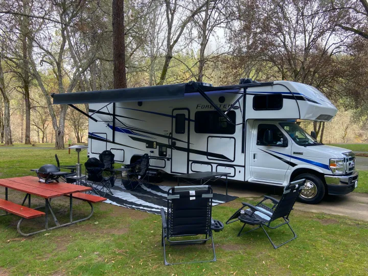 RV rental ready for adventure! This Class C motorhome is set up at a grassy campsite with an awning, chairs, grill, and picnic table.. Forest River Forester 2023