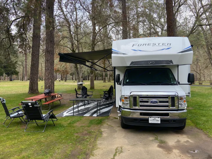 A white RV rental at a scenic campsite with an extended awning, outdoor living area, picnic table, and fire pit.. Forest River Forester 2023