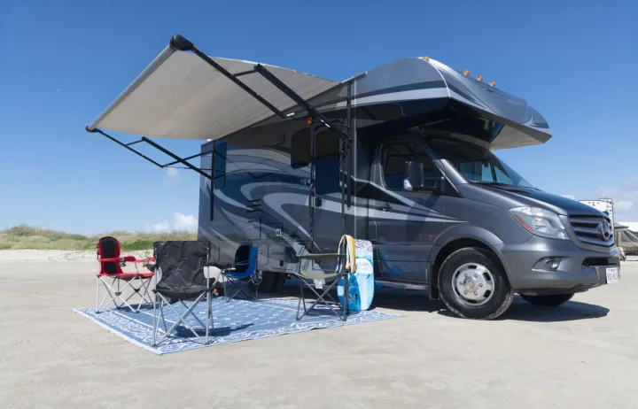 RV rental motorhome parked on a sandy area with an extended awning, outdoor rug, and chairs for comfortable camping.. Jayco Melbourne 2016