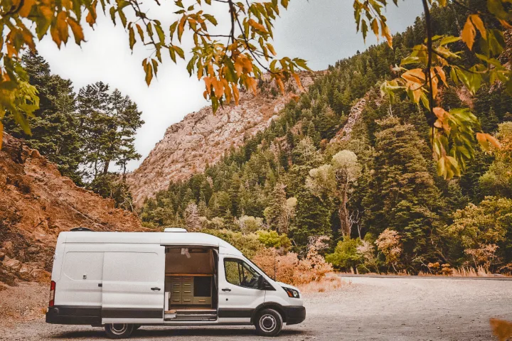 A white camper van, available for RV rental, parked on a dirt road amidst stunning mountain forests.. Ford Transit 2019