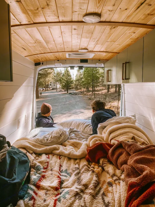 Inside an RV rental: Two people relax on a warm bed, admiring a wooded campground view from the open rear doors.. Ford Transit 2019