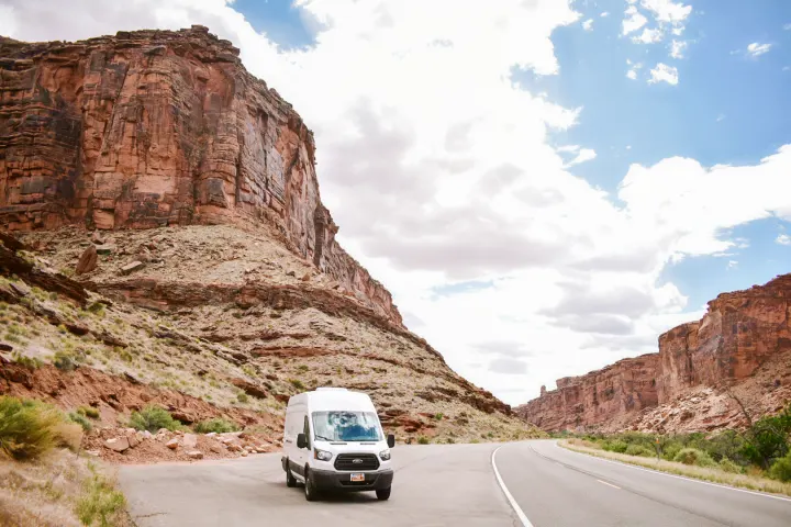 White Ford RV rental van on a scenic desert road, framed by towering red rock canyons under a cloudy sky.. Ford Transit 2019