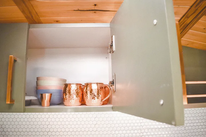 RV rental kitchen: open cabinet reveals stylish copper mugs, pastel bowls, and a small copper cup on hex tile.. Ford Transit 2019