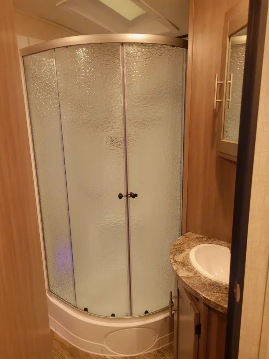This RV rental bathroom features a compact curved shower with textured glass doors and a corner vanity with a sink.. Crossroads Sunset Trail 2023
