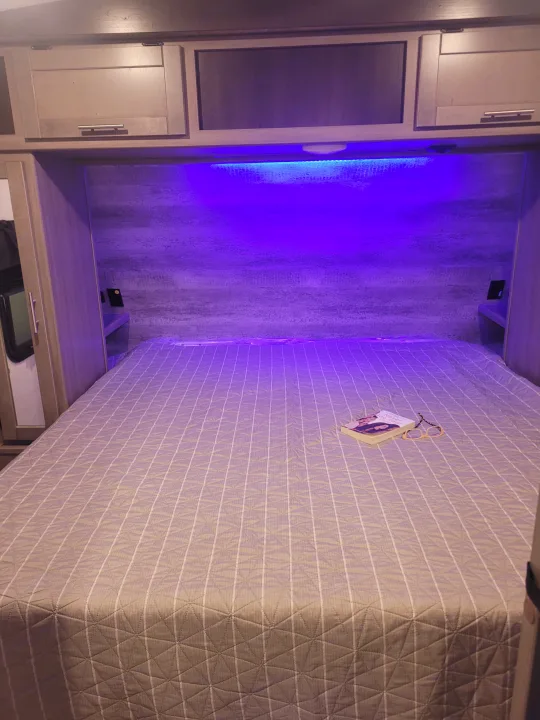 Experience comfort in this RV rental bedroom: illuminated by blue light, featuring storage, a cozy bed, book, and glasses.. Crossroads Sunset Trail 2023
