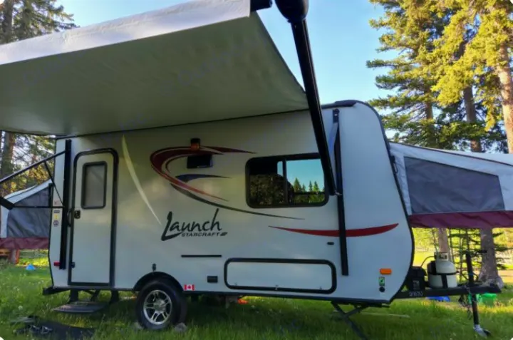 This grey Launch Starcraft hybrid travel trailer, ideal for an RV rental, is set up at a grassy campsite under an awning with trees.. Starcraft Launch 2016