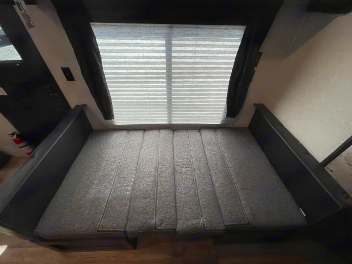 RV rental interior: A gray cushioned bed/dinette area under a window with blinds, flanked by dark curtains. Fire extinguisher on left.. Forest River Cherokee Grey Wolf Special Edition 2022
