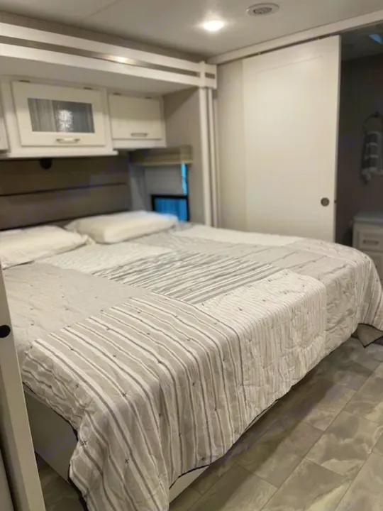Clean and modern RV rental bedroom featuring a comfy bed with a patterned quilt, overhead cabinets, and a sliding door.. Other Other 2021