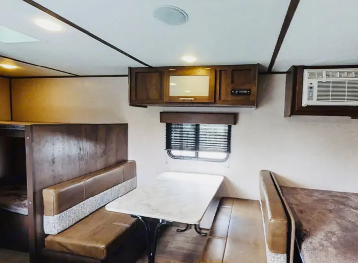 An RV rental interior showing a dinette with brown and grey upholstered seating, bunk beds, wood overhead cabinets, and an AC unit.. Coleman Other 2021
