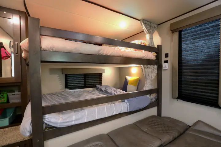 Comfortable bunk beds inside an RV rental with soft duvets, pillows, and a window with blinds. Shelf storage nearby.. Grand Design Transcend Xplor 2022