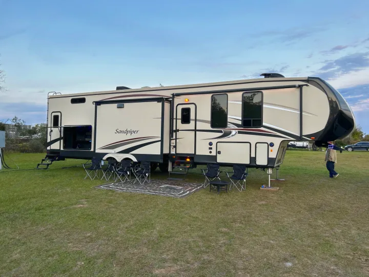 A large, light-colored fifth-wheel RV rental is fully set up on a grassy campsite with an outdoor seating area.. Forest River Sandpiper 2018