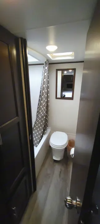 An RV rental bathroom featuring a white toilet, patterned shower curtain, mirror, and natural light from a window.. Jayco Jay Flight SLX 184 BS 2019
