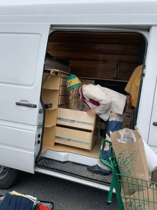Inside an RV rental, a person with a hat and fleece explores custom wood drawers and shelves in their mobile home.. Mercedes Sprinter 2006