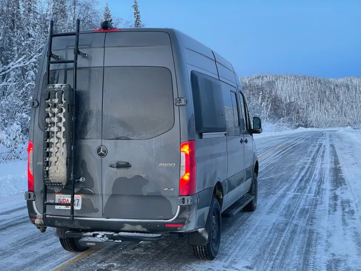 An RV rental, a gray Mercedes Sprinter van, parked on an icy Alaska road amid snowy trees, ready for winter adventure.. Mercedes Sprinter 2022