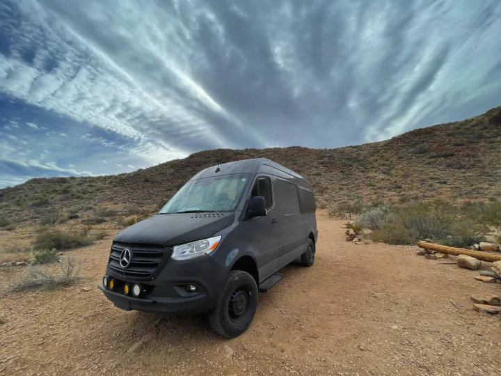 Experience desert adventures! A dark gray Sprinter camper van, perfect for RV rental, parked on a dusty trail with dramatic clouds.. Mercedes Sprinter 2022