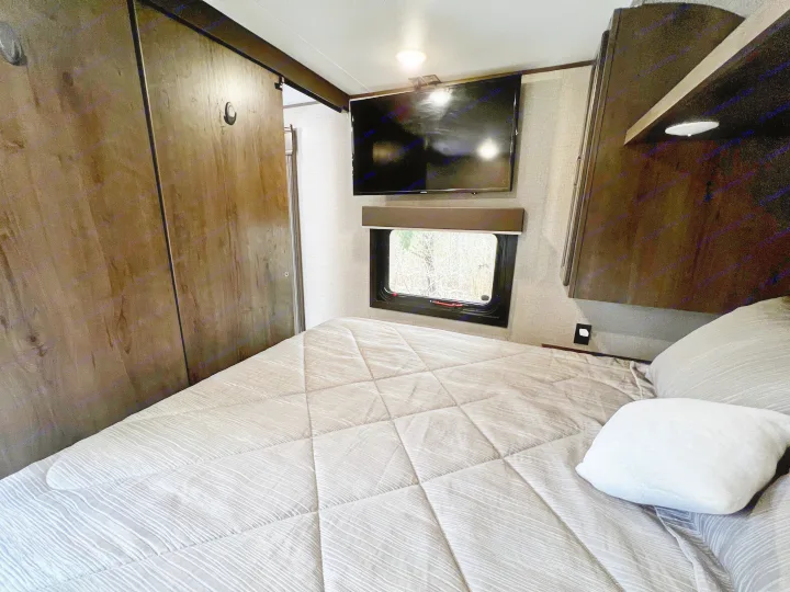 View inside an RV rental bedroom, showing a bed with patterned comforter, wall-mounted TV, and window with outdoor view.. Jayco Jay Flight SLX 2020