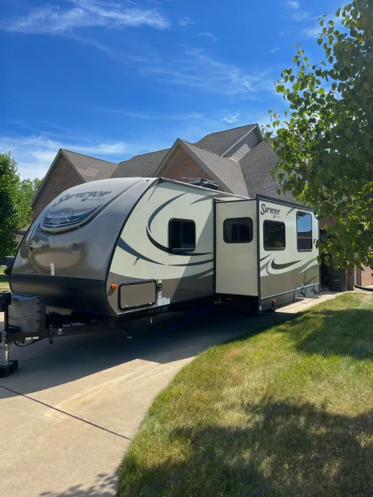 A large tan travel trailer RV rental sits in a driveway, its slide-out extended, under a bright blue sky, ready for travel.. Forest River Surveyor 2016