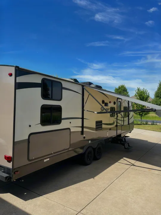 Modern tan and brown travel trailer with extended awning parked on a driveway. Consider an RV rental for your next road trip!. Forest River Surveyor 2016