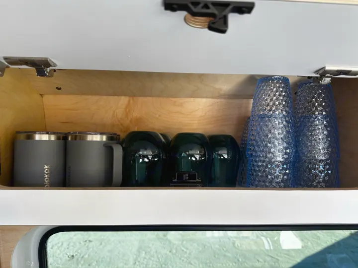 Inside an RV rental kitchen cabinet: neatly organized gray mugs, dark green glasses, and textured blue cups.. VanCraft VanCraft Class B 2022