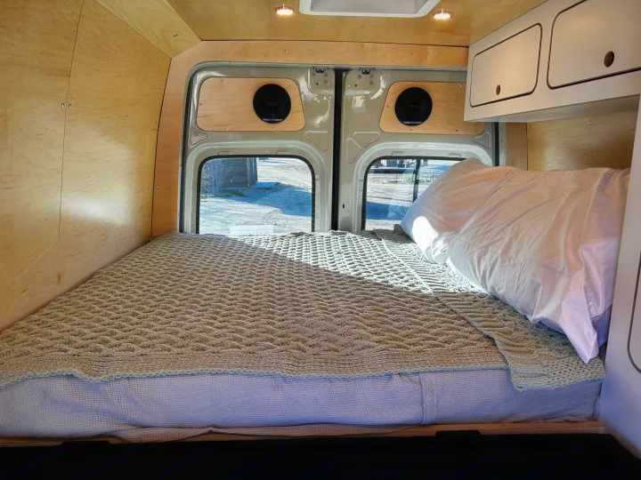 Comfortable RV rental camper van bed with a knitted blanket, wood-paneled interior, and rear windows.. VanCraft VanCraft Class B 2022