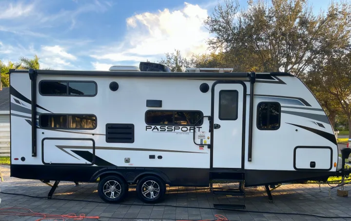Side view of a white "Passport" travel trailer RV rental, parked on pavers with trees & sky. Perfect for adventures.. Keystone RV Passport SL 2022
