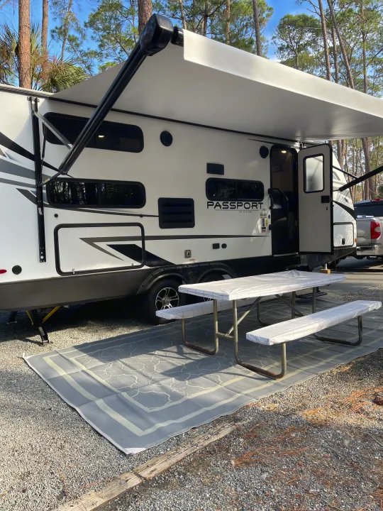 Enjoying an RV rental at a gravel campsite, complete with a white travel trailer, extended awning, picnic table & outdoor rug.. Keystone RV Passport SL 2022