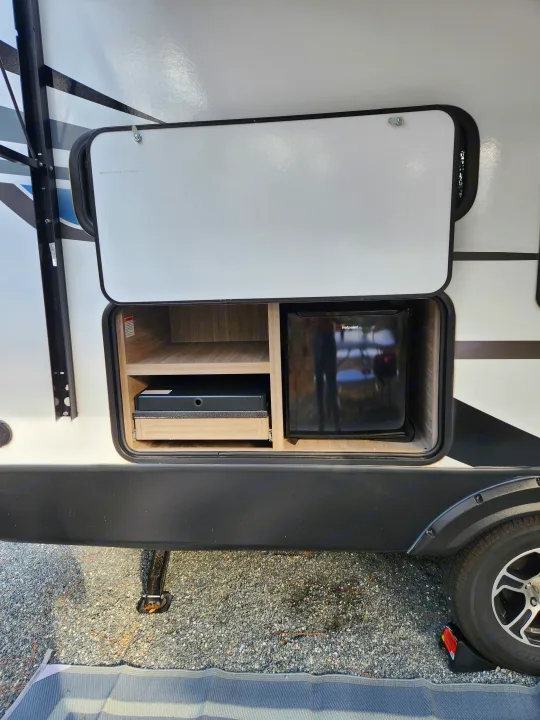 Open outdoor kitchen of an **RV rental**, featuring a compact fridge, shelves, and a gray mat on gravel.. Keystone RV Passport SL 2022