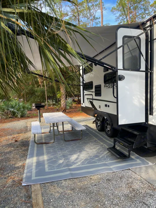 RV rental campsite setup: white RV with extended awning, outdoor rug, covered picnic table, and grill.. Keystone RV Passport SL 2022