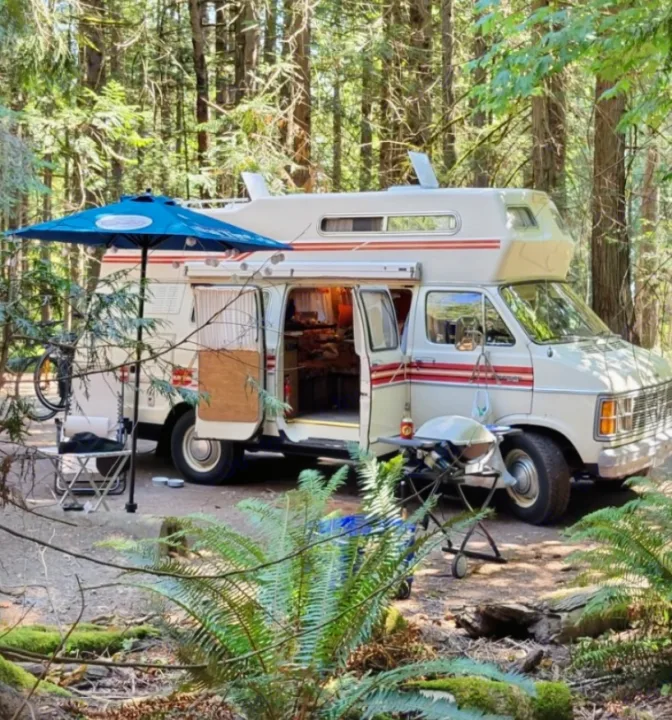 An RV rental provides adventure! Vintage camper van at a serene forest campsite with umbrella, grill, and chair setup.. Frontier Other 1979